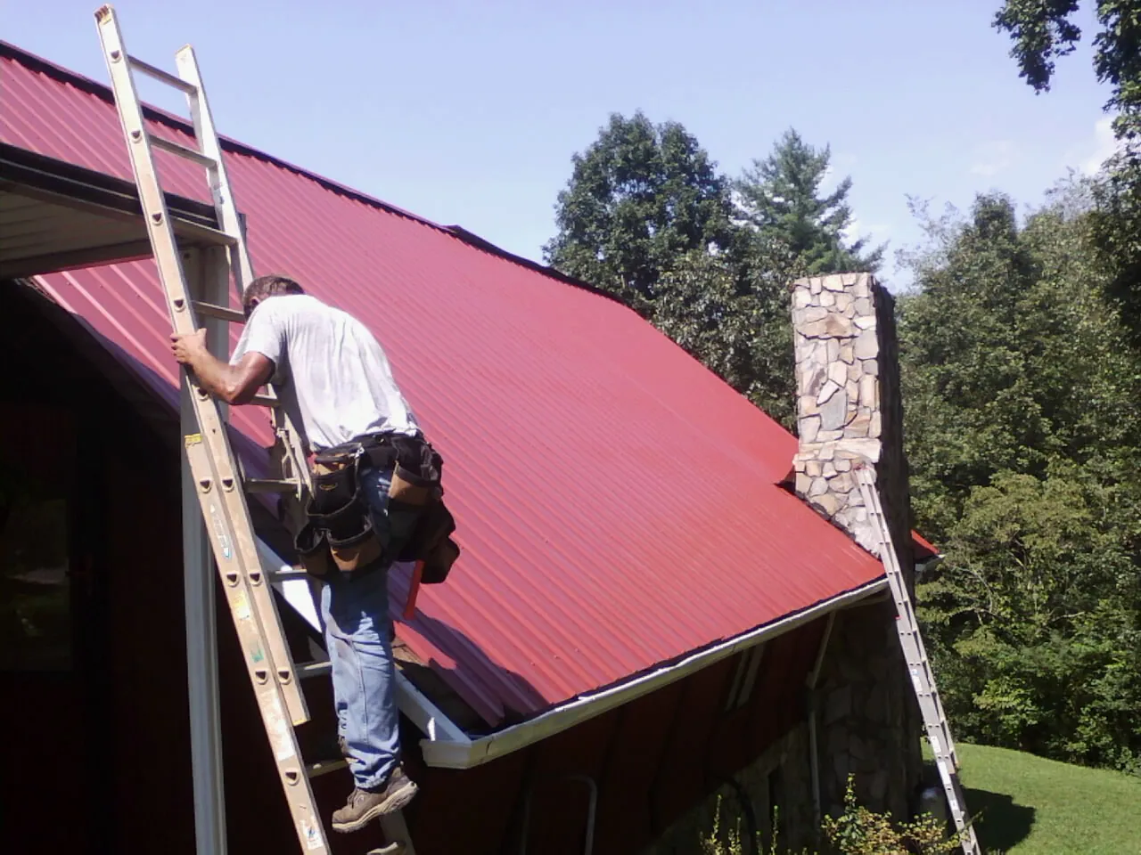 Roofer on a ladder beside a red metal roof and stone chimney — Soffit & Fascia Repair by Roofing Brothers in Intercourse